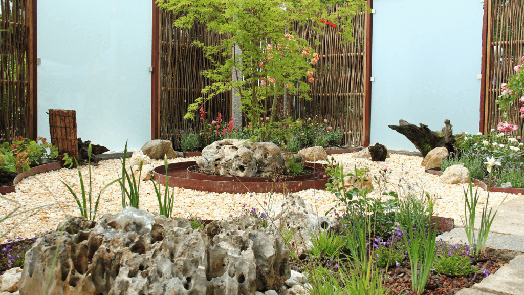 The example features a sensory garden with a fountain, flower beds, and various outdoor plants and flowers. The landscaping is designed to create a peaceful and sensory-rich environment, enhanced by the mix of textures in the fence, flower bed borders and gravel pathway.